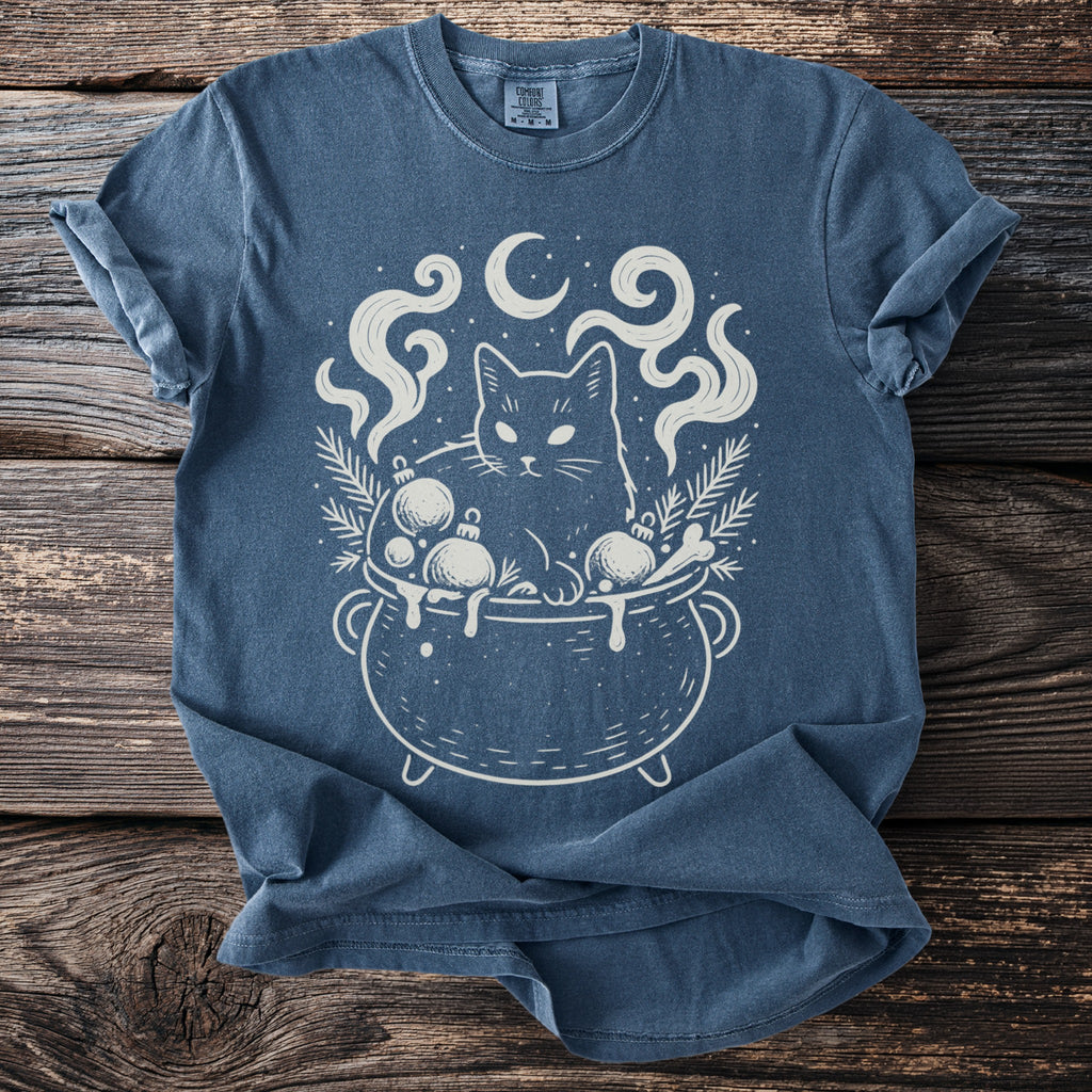 Blue t-shirt with a white graphic of a cat in a cauldron with Christmas ornaments and moon.