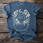 Blue t-shirt with a white graphic of a cat in a cauldron with Christmas ornaments and moon.