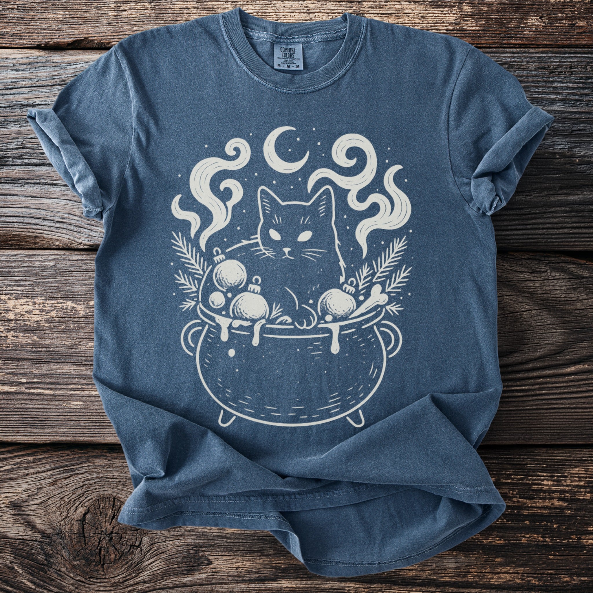 Blue t-shirt with a white graphic of a cat in a cauldron with Christmas ornaments and moon.
