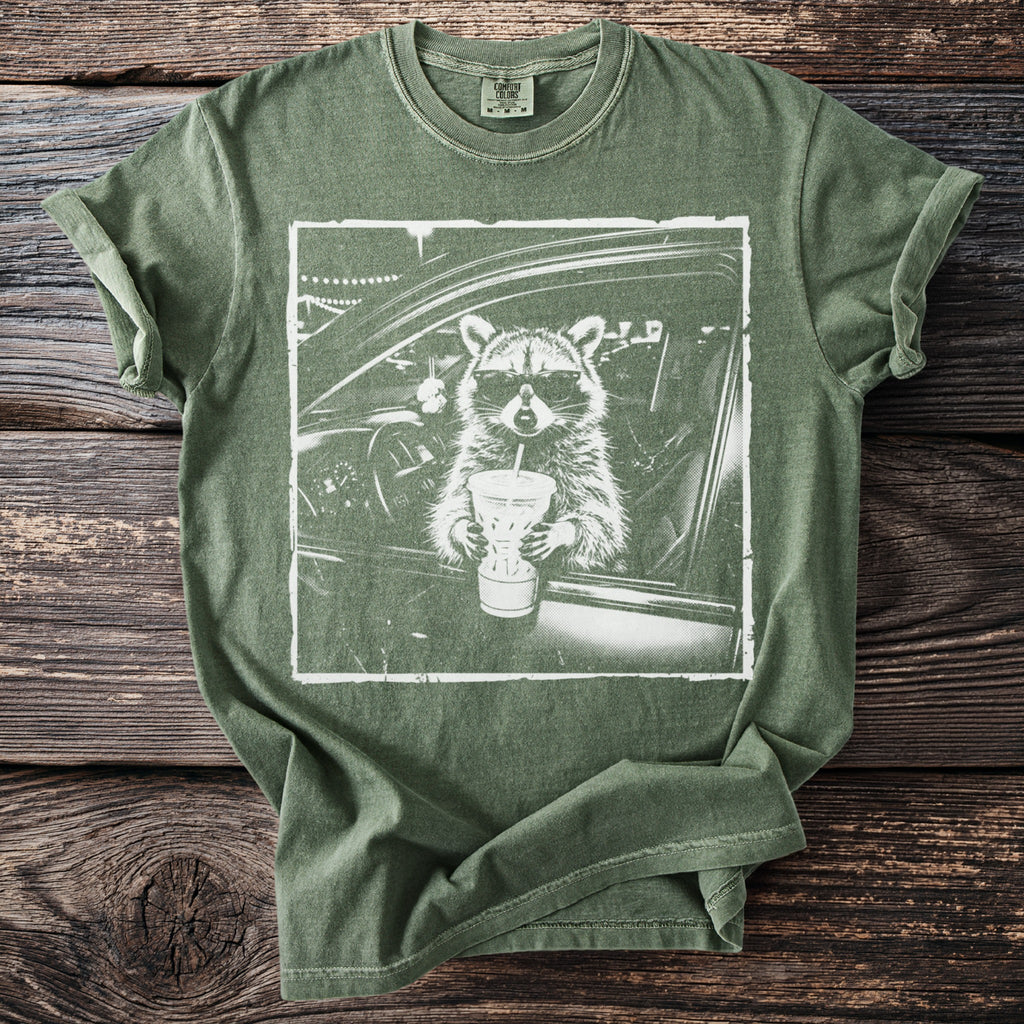 Green t-shirt with a graphic of a raccoon wearing sunglasses driving a car and holding a drink.