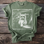 Green t-shirt with a graphic of a raccoon wearing sunglasses driving a car and holding a drink.