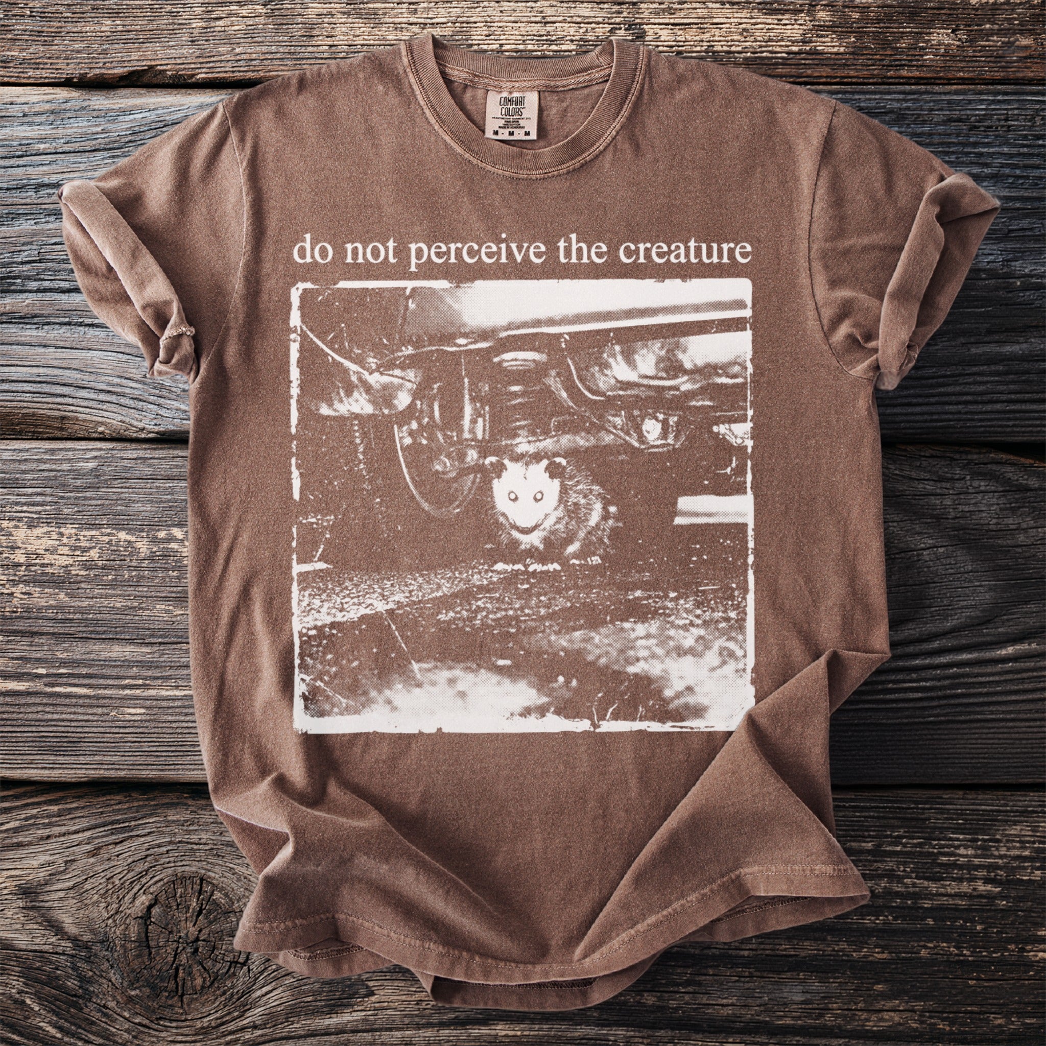 Brown t-shirt with possum under a car and text "do not perceive the creature".