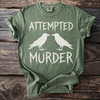 Green t-shirt with white text "ATTEMPTED MURDER" and two birds.