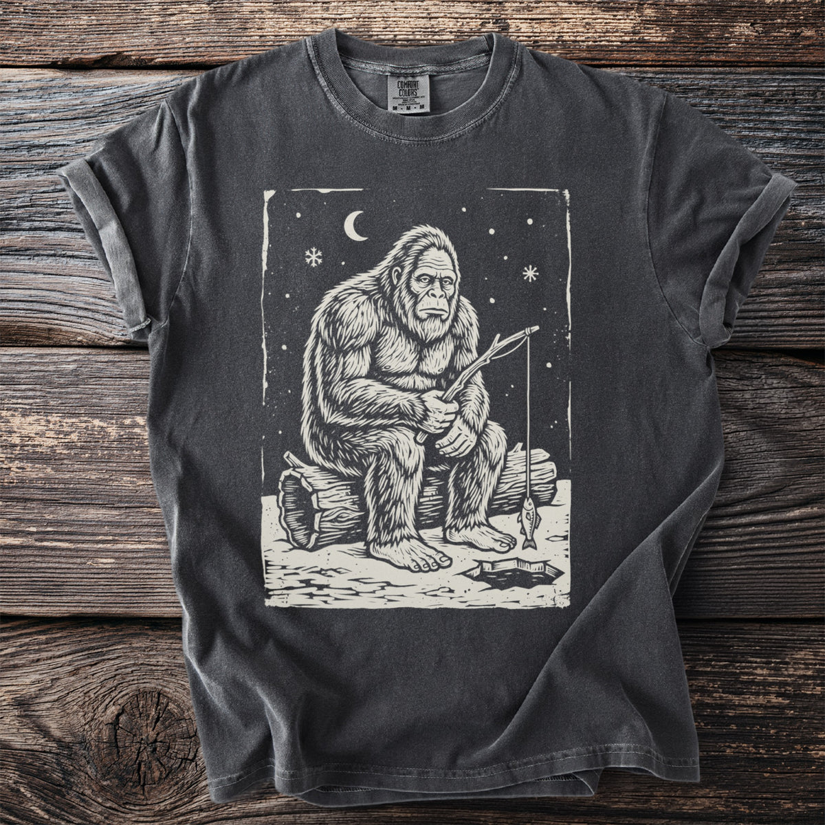 Pepper gray t-shirt with a cream folk-cut print: Bigfoot sits on a log with a stick rod over a hole in the ice, one small fish on the line.