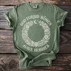 Green t-shirt with white text "GOD FORBID WOMEN HAVE HOBBIES" and floral wreath.