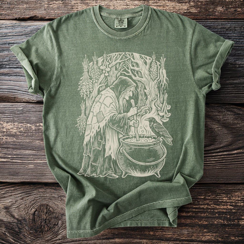 Green t-shirt with a white line drawing of a witch by a cauldron with a raven.