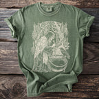 Green t-shirt with a white line drawing of a witch by a cauldron with a raven.