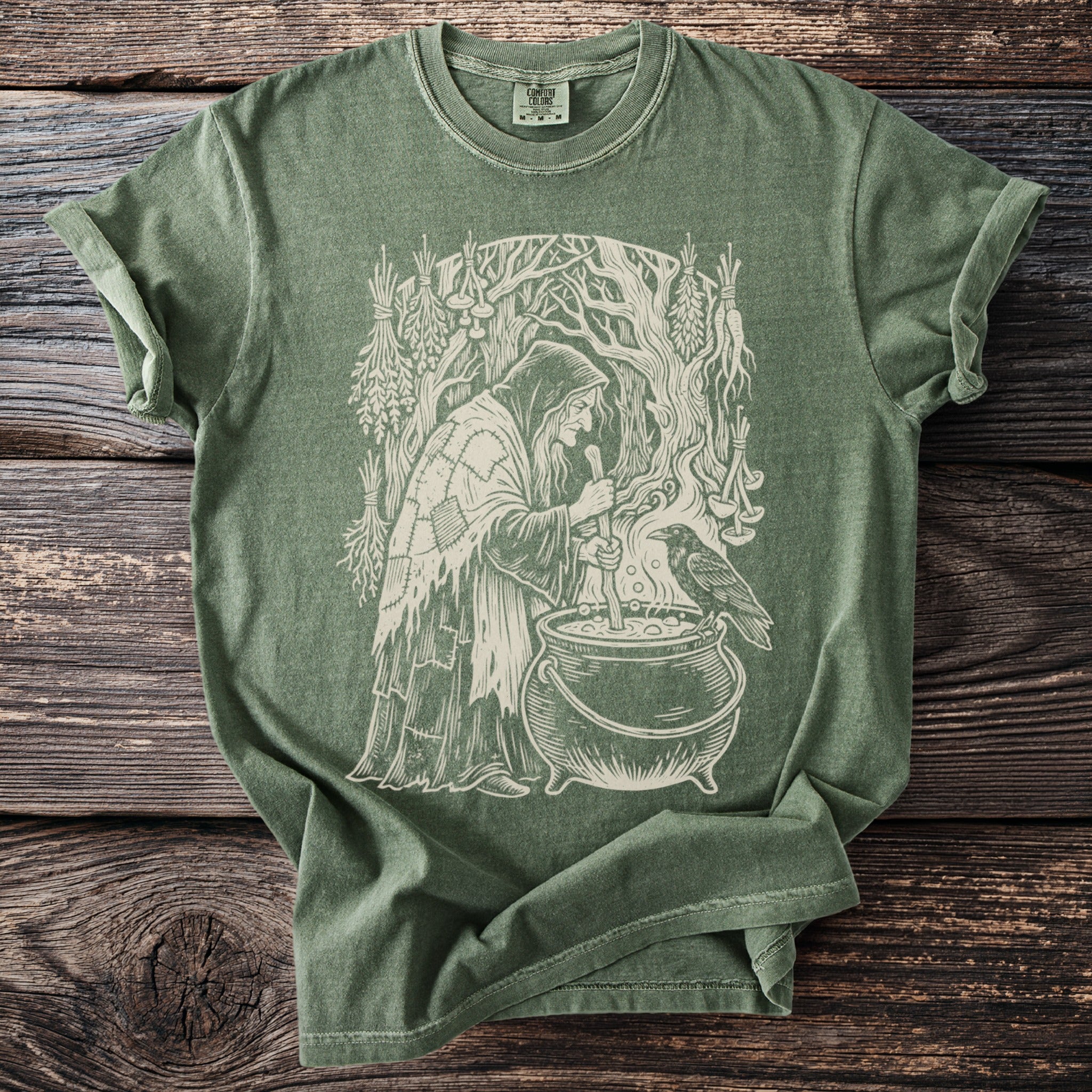 Green t-shirt with a white line drawing of a witch by a cauldron with a raven.