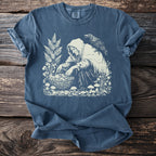 Graphic of hooded figure gathering mushrooms with a crow on shoulder, on a blue t-shirt.