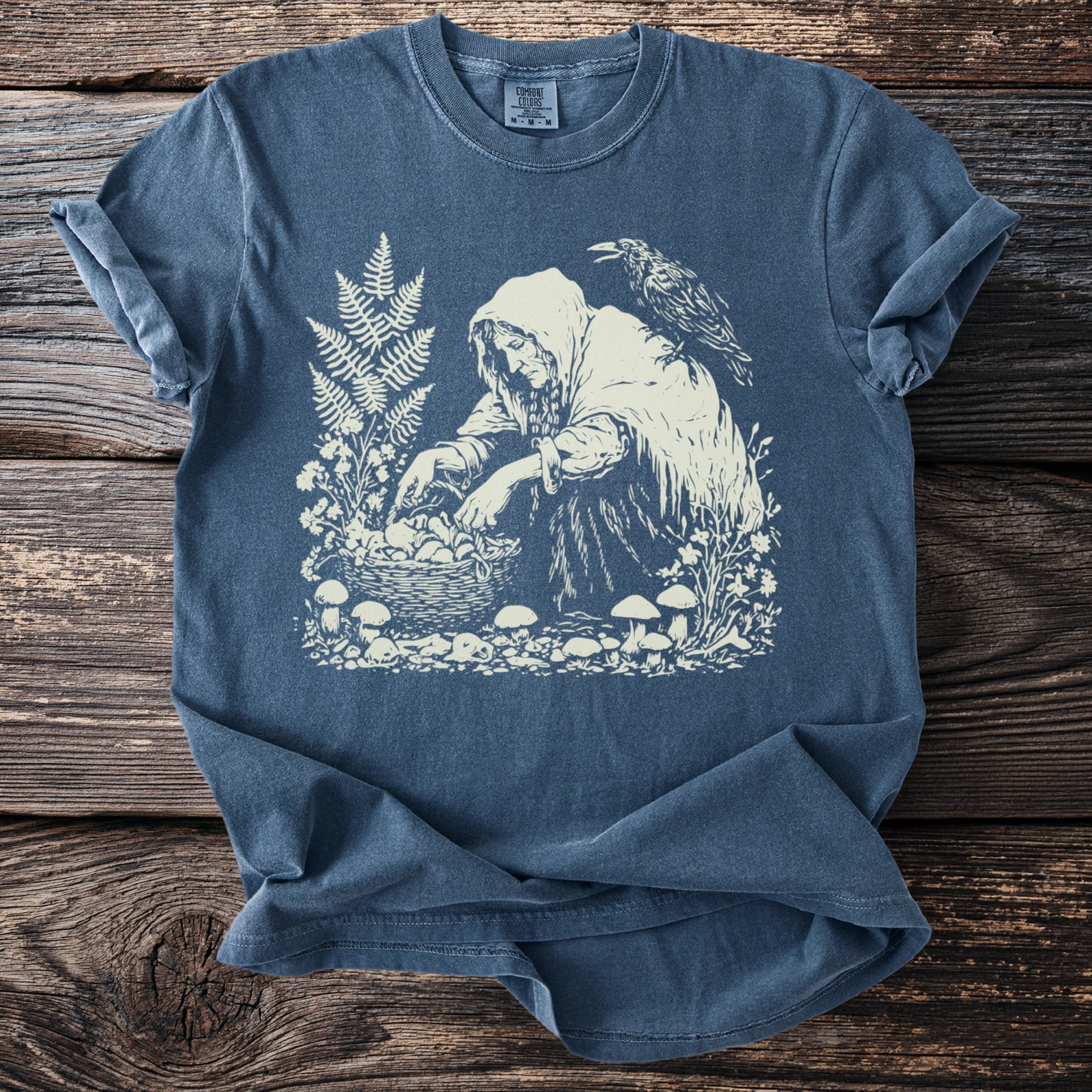 Graphic of hooded figure gathering mushrooms with a crow on shoulder, on a blue t-shirt.