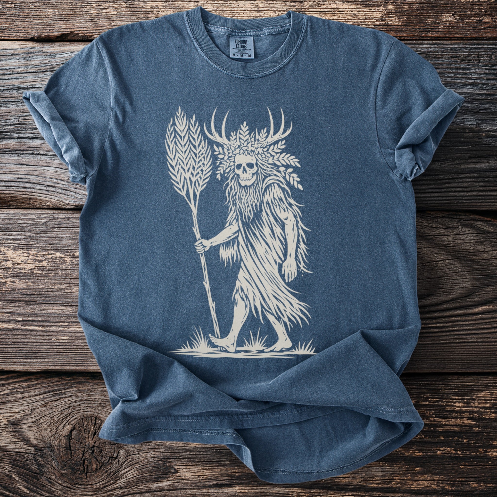 Blue t-shirt with a beige print of a skull-headed figure with antlers holding a staff.