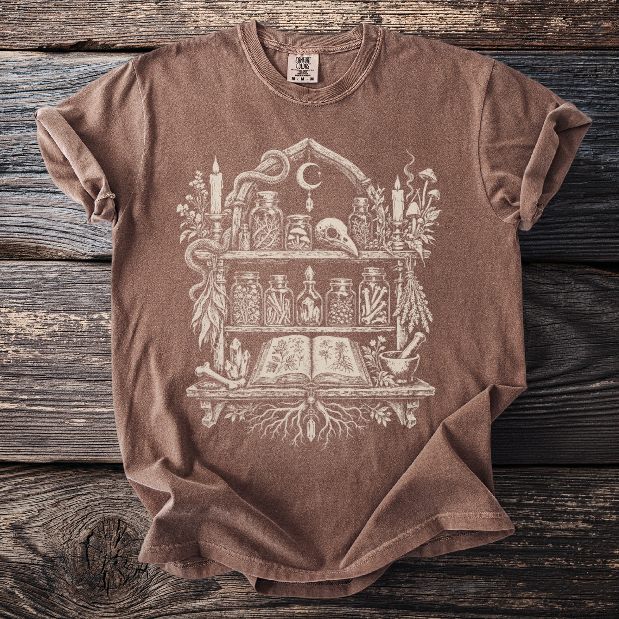 Brown t-shirt with an illustration of a witch's shelf filled with potions and curiosities.