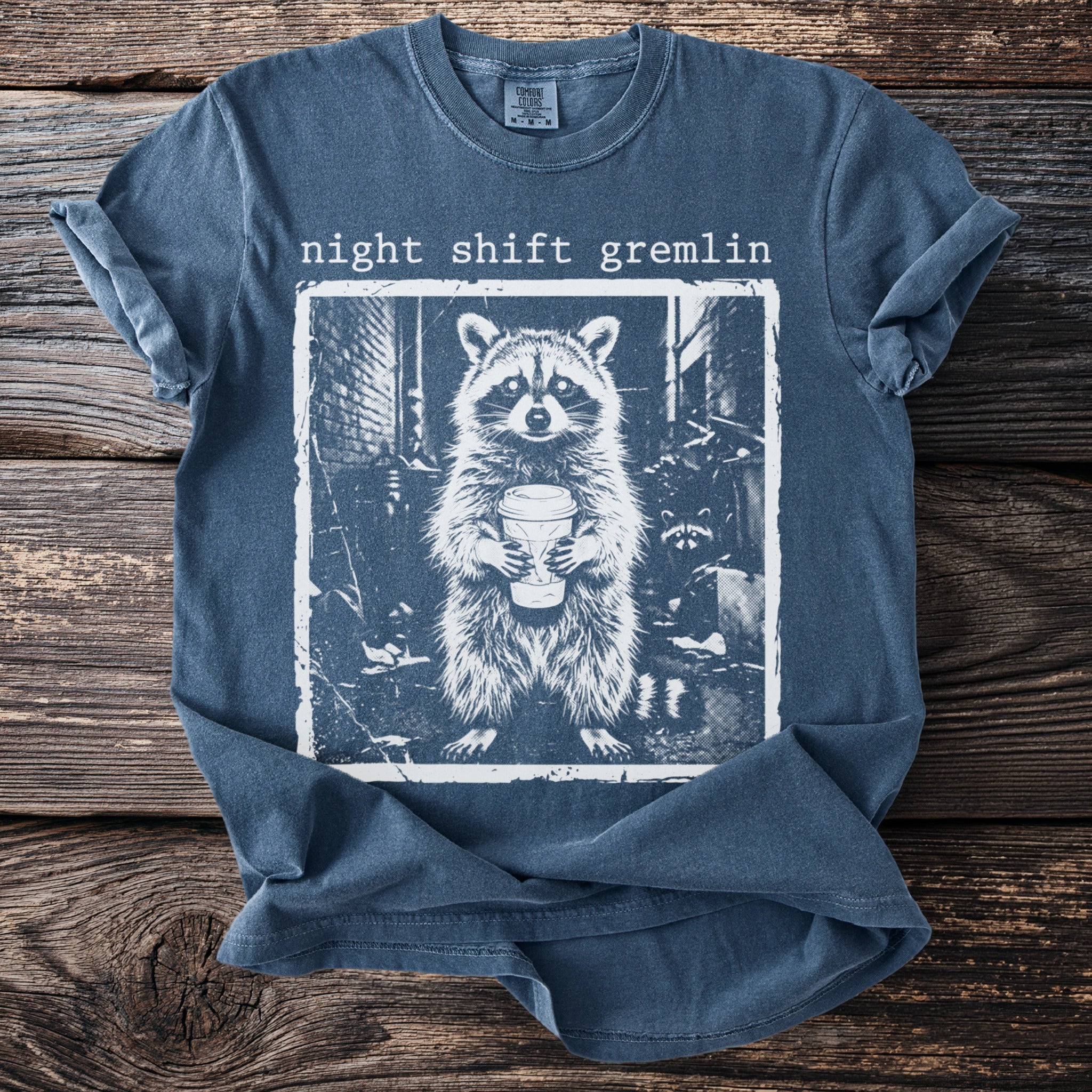 Blue t-shirt with "night shift gremlin" text and a raccoon holding a coffee cup graphic.