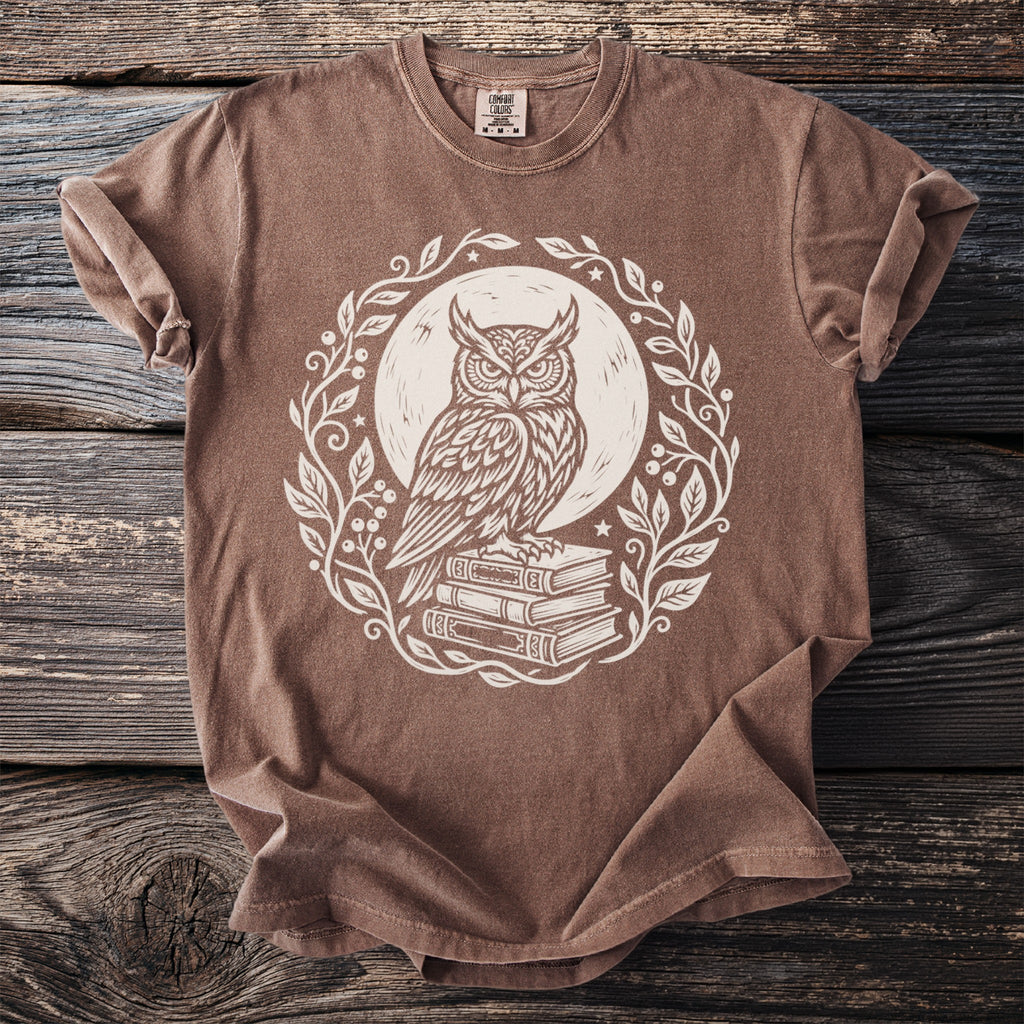 Brown t-shirt with a white illustration of an owl on stacked books within a leafy wreath.