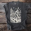 Pepper gray t-shirt with a cream folk-cut print: Bigfoot walks past a snowy cabin dragging a whole fir tree, big footprints behind him.
