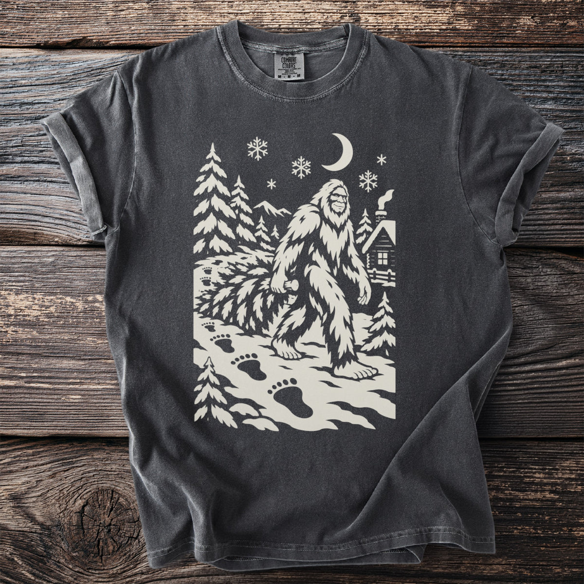 Pepper gray t-shirt with a cream folk-cut print: Bigfoot walks past a snowy cabin dragging a whole fir tree, big footprints behind him.