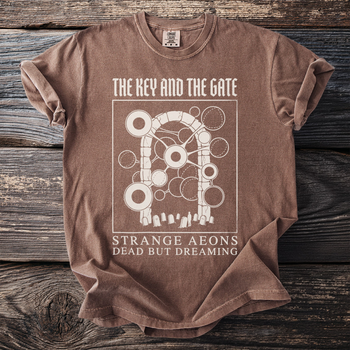 Brown t-shirt with "The Key and the Gate" graphic and text "Strange Aeons Dead But Dreaming".