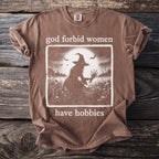 Brown t-shirt with graphic of woman in witch hat and text "god forbid women have hobbies."