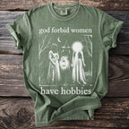 Olive green t-shirt with "god forbid women have hobbies" text and witchy graphic.