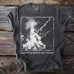 Dark gray t-shirt with graphic of Bigfoot grabbing a hot dog over a campfire and text.