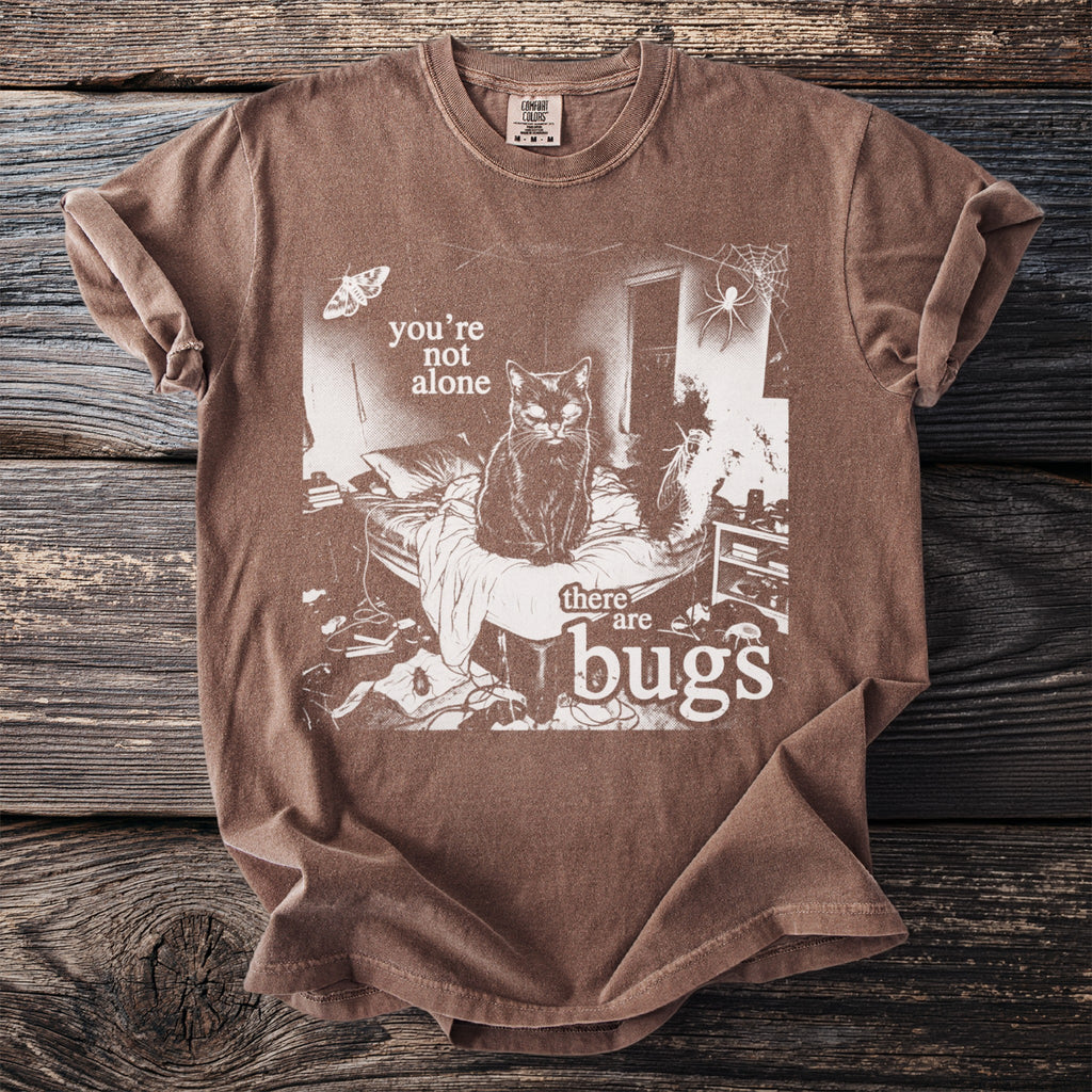 Brown t-shirt with white graphic of a cat on a bed, moth, spider, and text "you're not alone there are bugs".