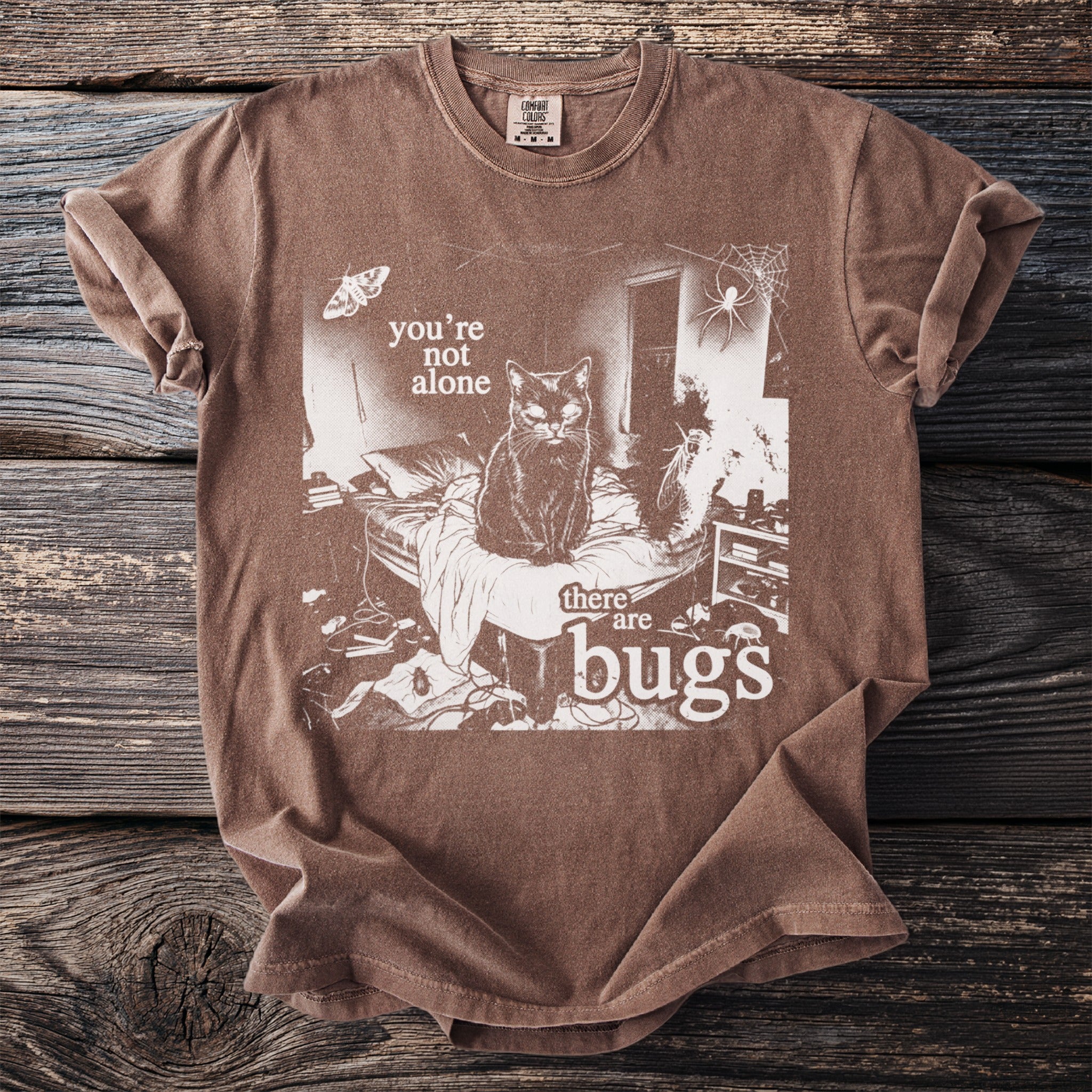 Brown t-shirt with white graphic of a cat on a bed, moth, spider, and text "you're not alone there are bugs".