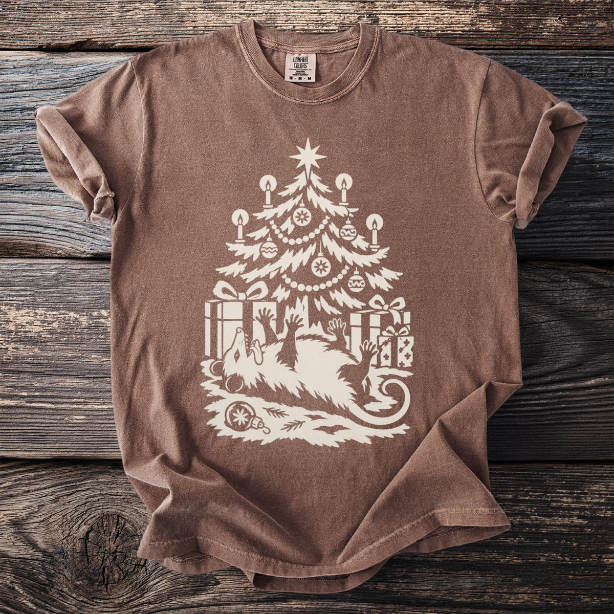 Espresso brown t-shirt with a cream folk-cut print: an opossum lies on its back under a candlelit Christmas tree, playing dead among the presents.