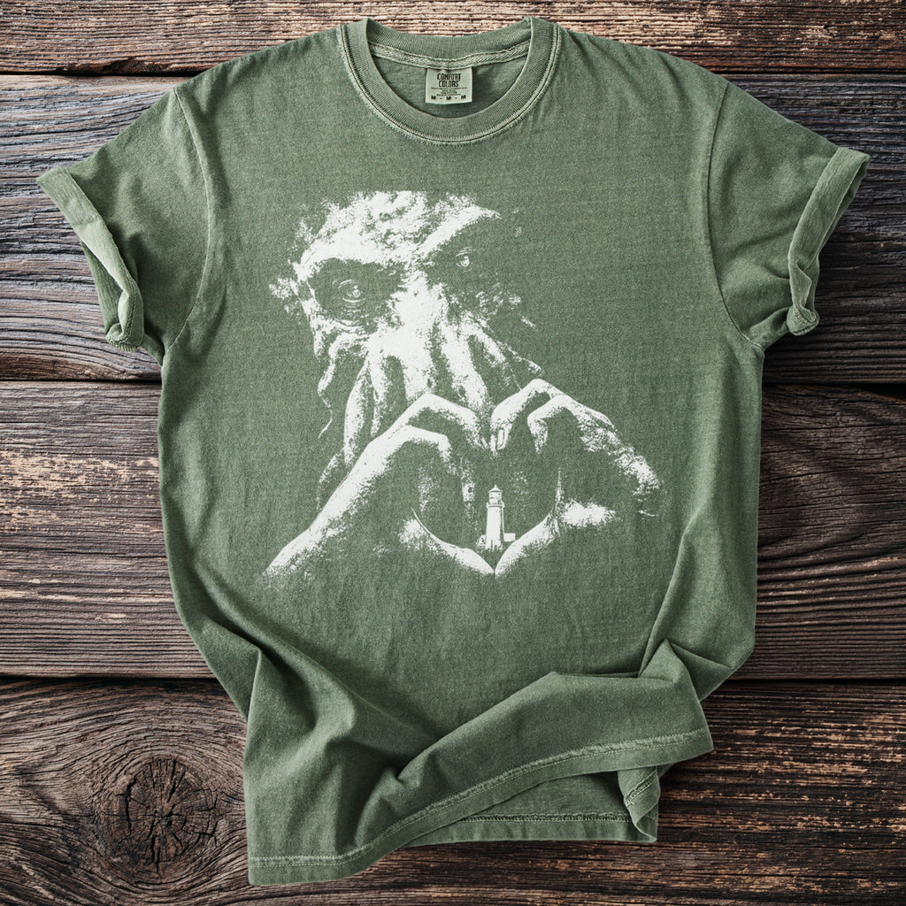 Green t-shirt with white abstract graphic of creature and heart-shaped hands holding a lighthouse.