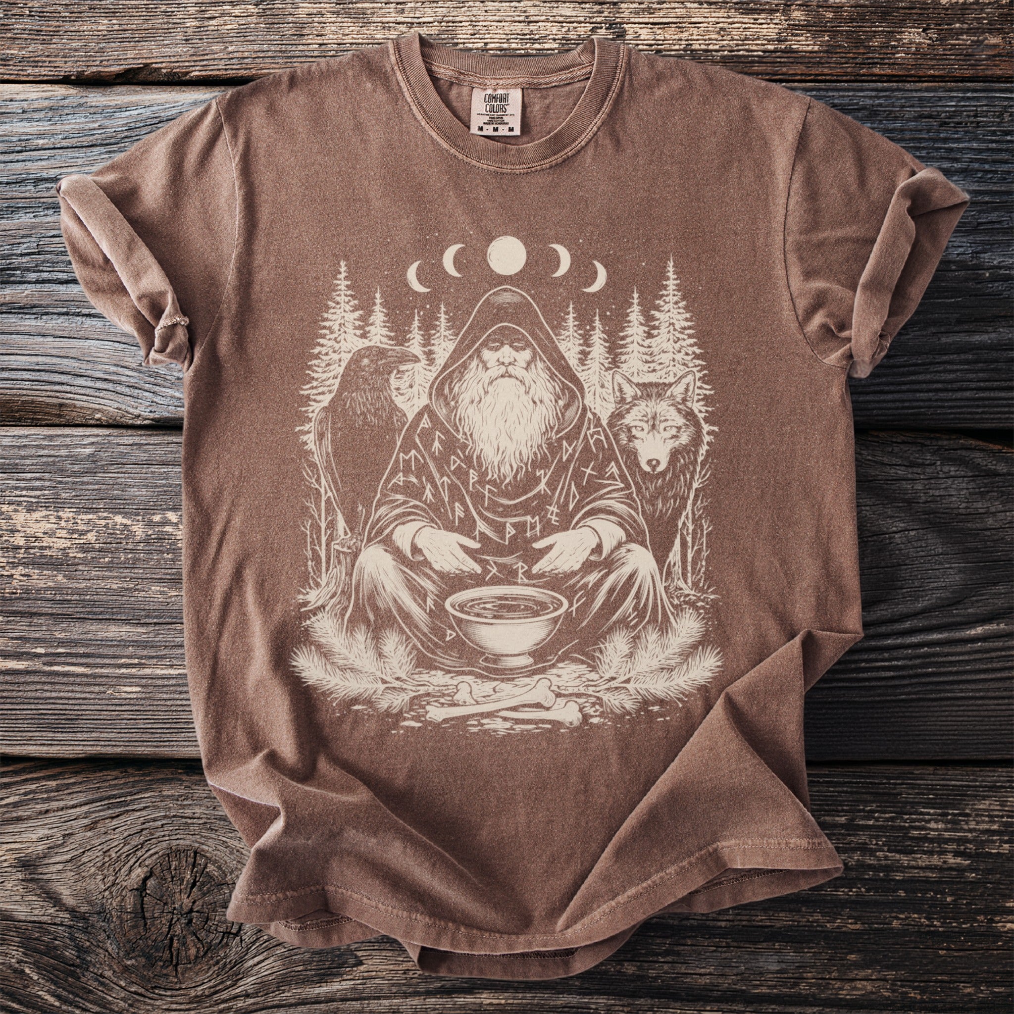 Brown t-shirt with a white graphic of a hooded figure with a beard, raven, wolf, trees, and moon phases.