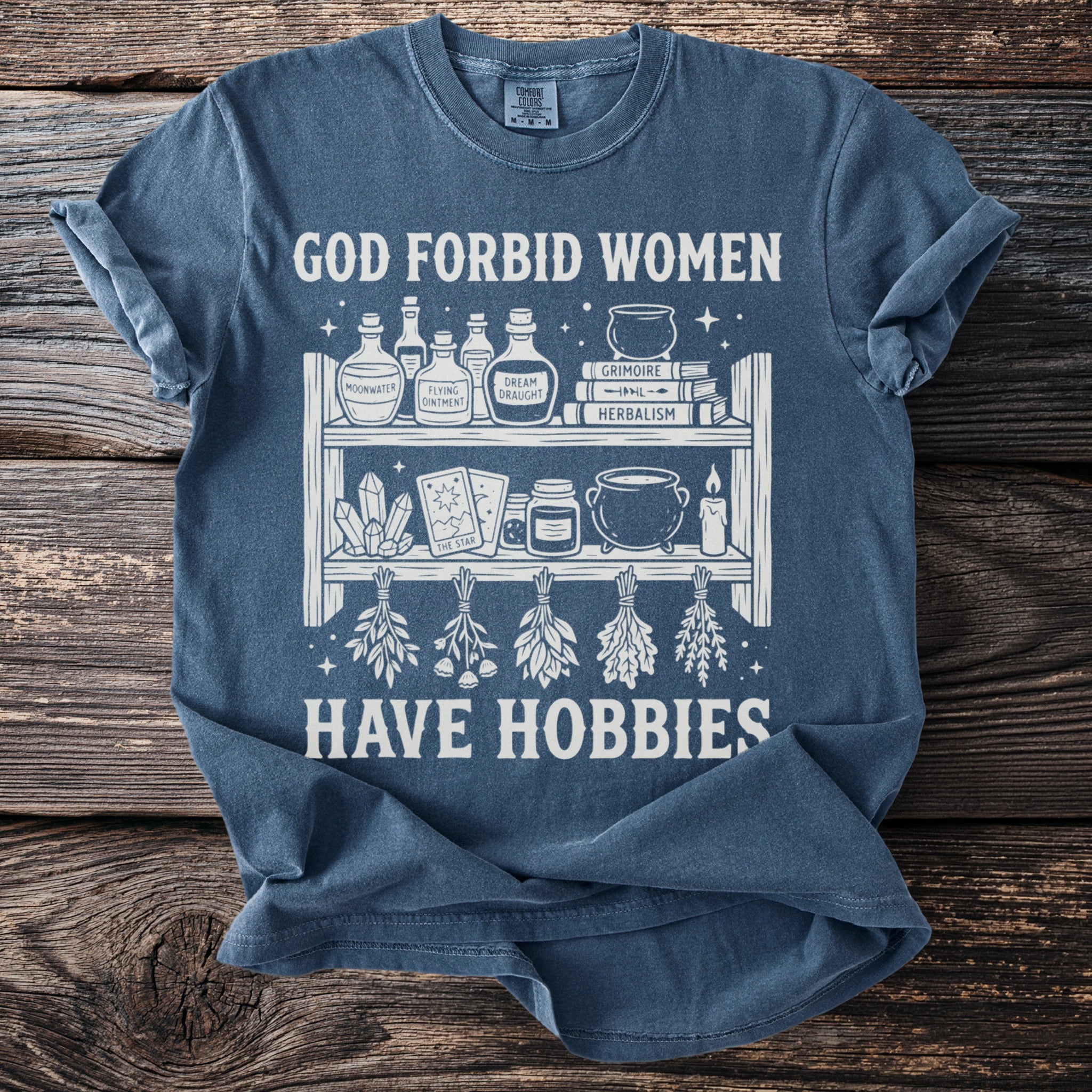 Blue t-shirt with "God forbid women have hobbies" and witchy items graphic.