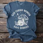 Blue t-shirt with white graphic of a frog, mushroom, moon, and stars with text "God Forbid Women Have Hobbies".