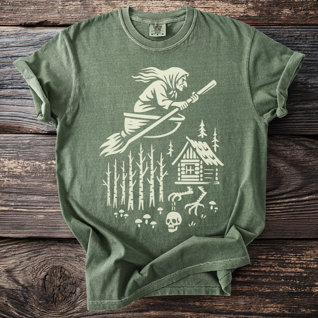 Green t-shirt with white graphic of a witch on a broomstick, cabin, and skull.
