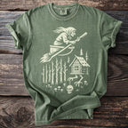 Green t-shirt with white graphic of a witch on a broomstick, cabin, and skull.