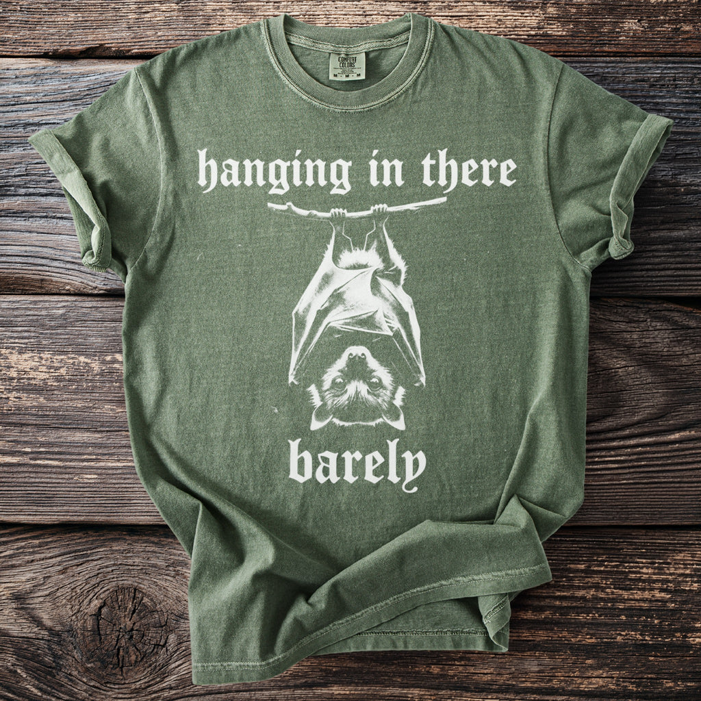 Green t-shirt with white text and a bat graphic reading "hanging in there barely".