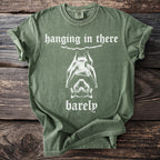 Green t-shirt with white text and a bat graphic reading "hanging in there barely".