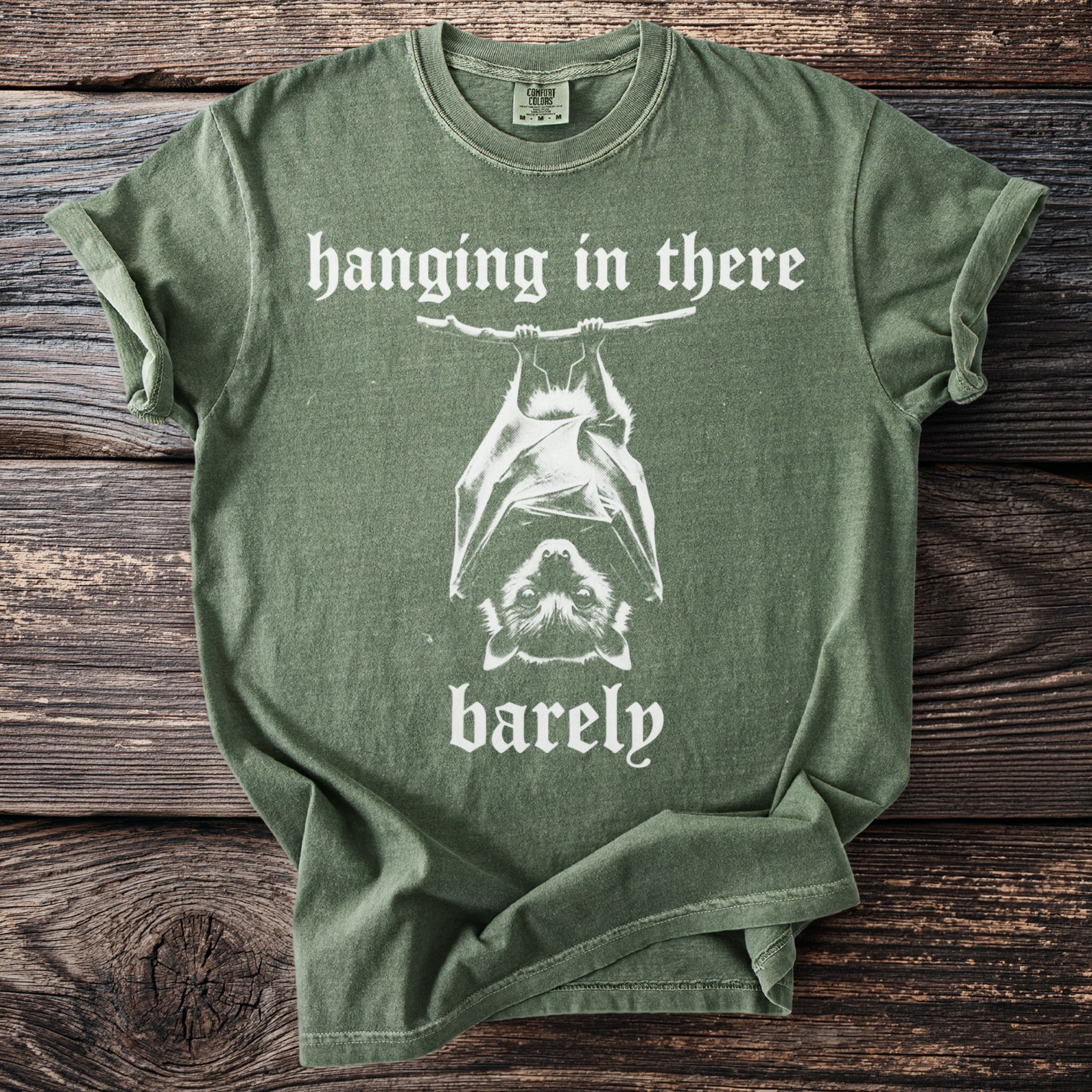 Green t-shirt with white text and a bat graphic reading "hanging in there barely".