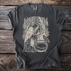 Dark grey t-shirt with a cream linocut-style design of a witch stirring a cauldron with a raven watching.