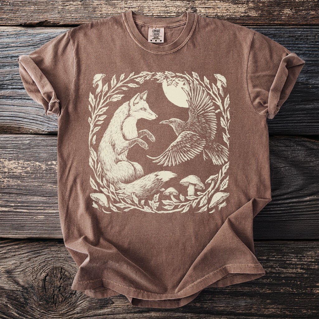 Brown t-shirt with a cream illustration of a fox and raven in a forest clearing.