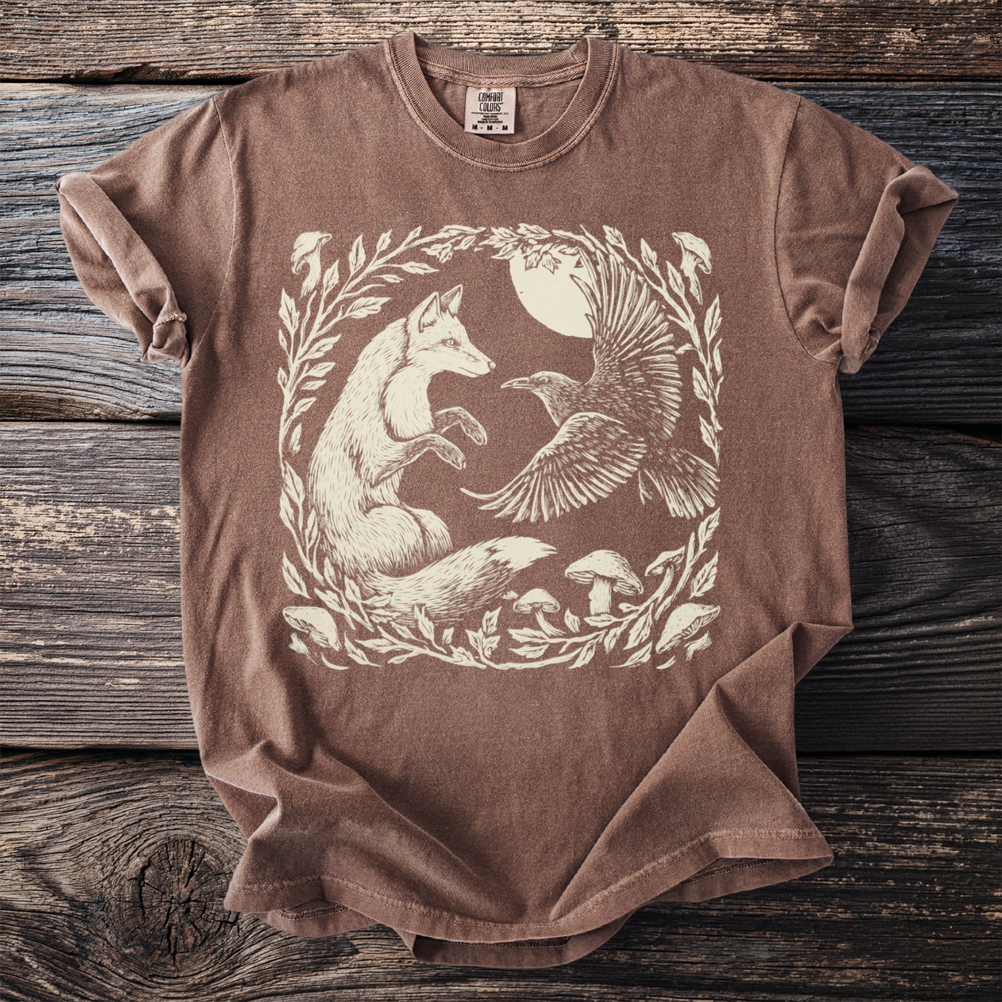 Brown t-shirt with a cream illustration of a fox and raven in a forest clearing.