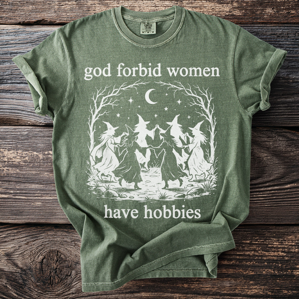 Green t-shirt with white graphic of witches dancing under a moon and text "god forbid women have hobbies".