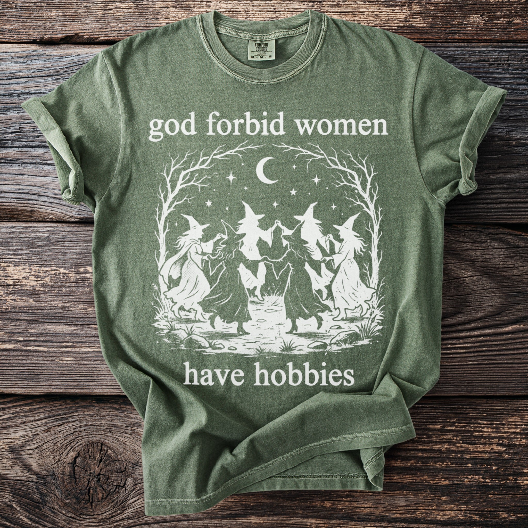 Green t-shirt with white graphic of witches dancing under a moon and text "god forbid women have hobbies".