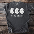 Three white ghosts in Santa hats, one holding a skull, on a dark gray t-shirt with "Spooky & Bright" text.