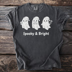Three white ghosts in Santa hats, one holding a skull, on a dark gray t-shirt with "Spooky & Bright" text.