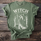 Green t-shirt with white graphic of a witch, cauldron, cat, trees, and stars.
