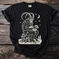 Black t-shirt with a cream folk-cut print: Krampus sits on a stump in the snow, carefully tying a small present with ribbon.