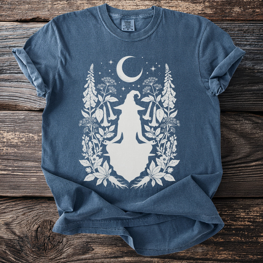 Blue t-shirt with a white graphic of a seated witch and crescent moon with botanical elements.