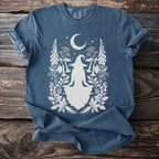 Blue t-shirt with a white graphic of a seated witch and crescent moon with botanical elements.