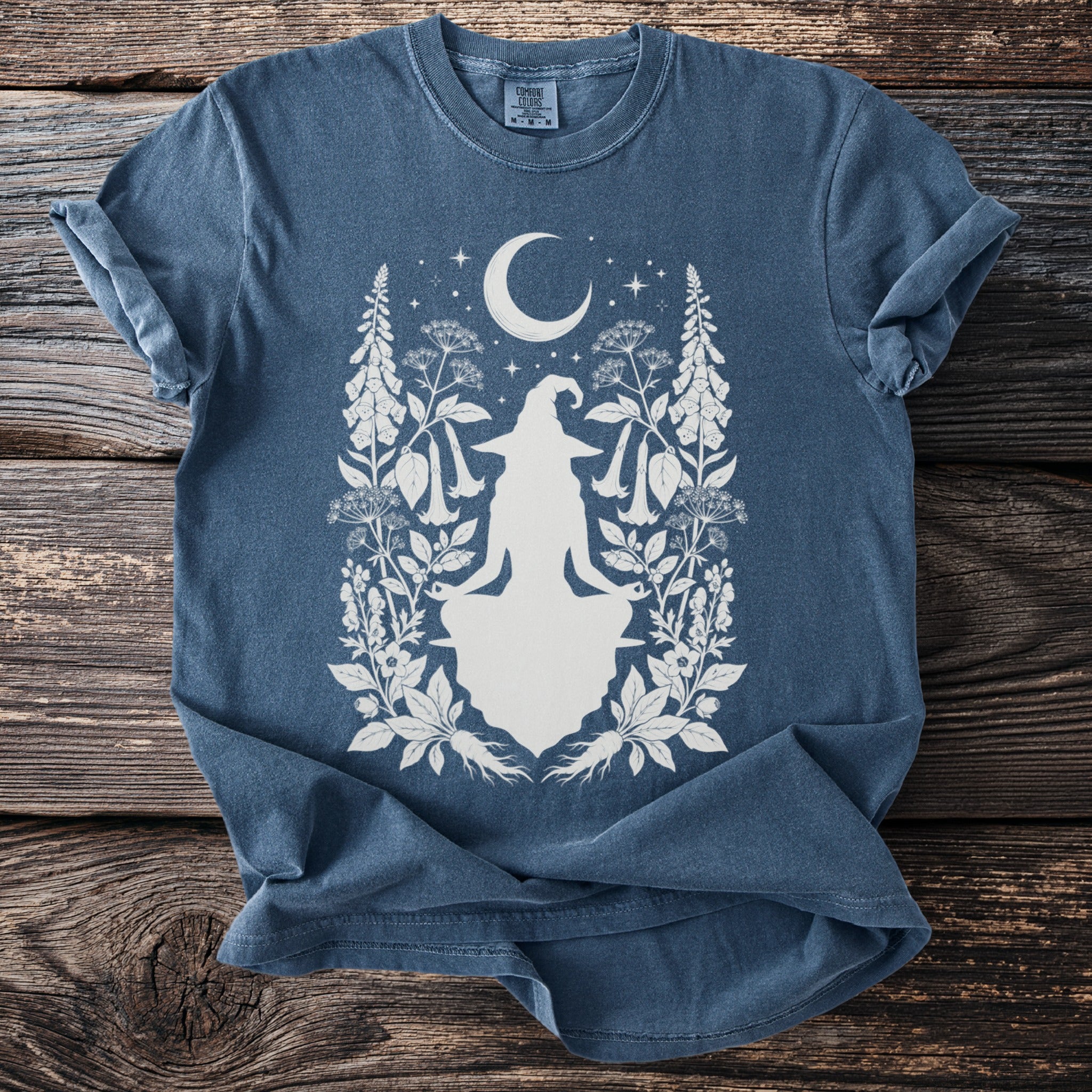 Blue t-shirt with a white graphic of a seated witch and crescent moon with botanical elements.