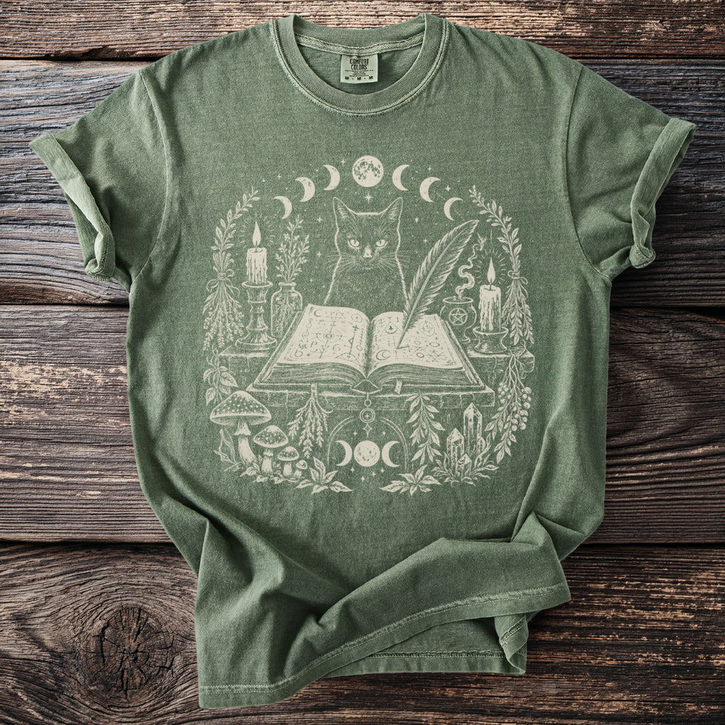 Green t-shirt with a cream graphic of a cat, open book, candles, and botanical elements.