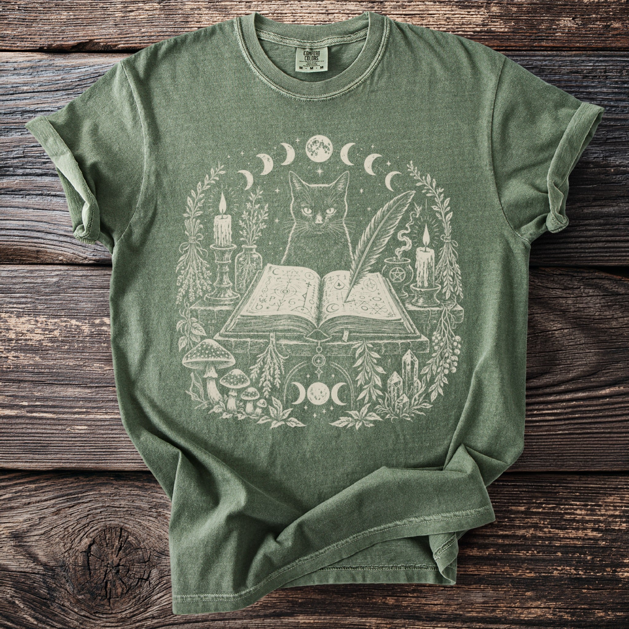 Green t-shirt with a cream graphic of a cat, open book, candles, and botanical elements.